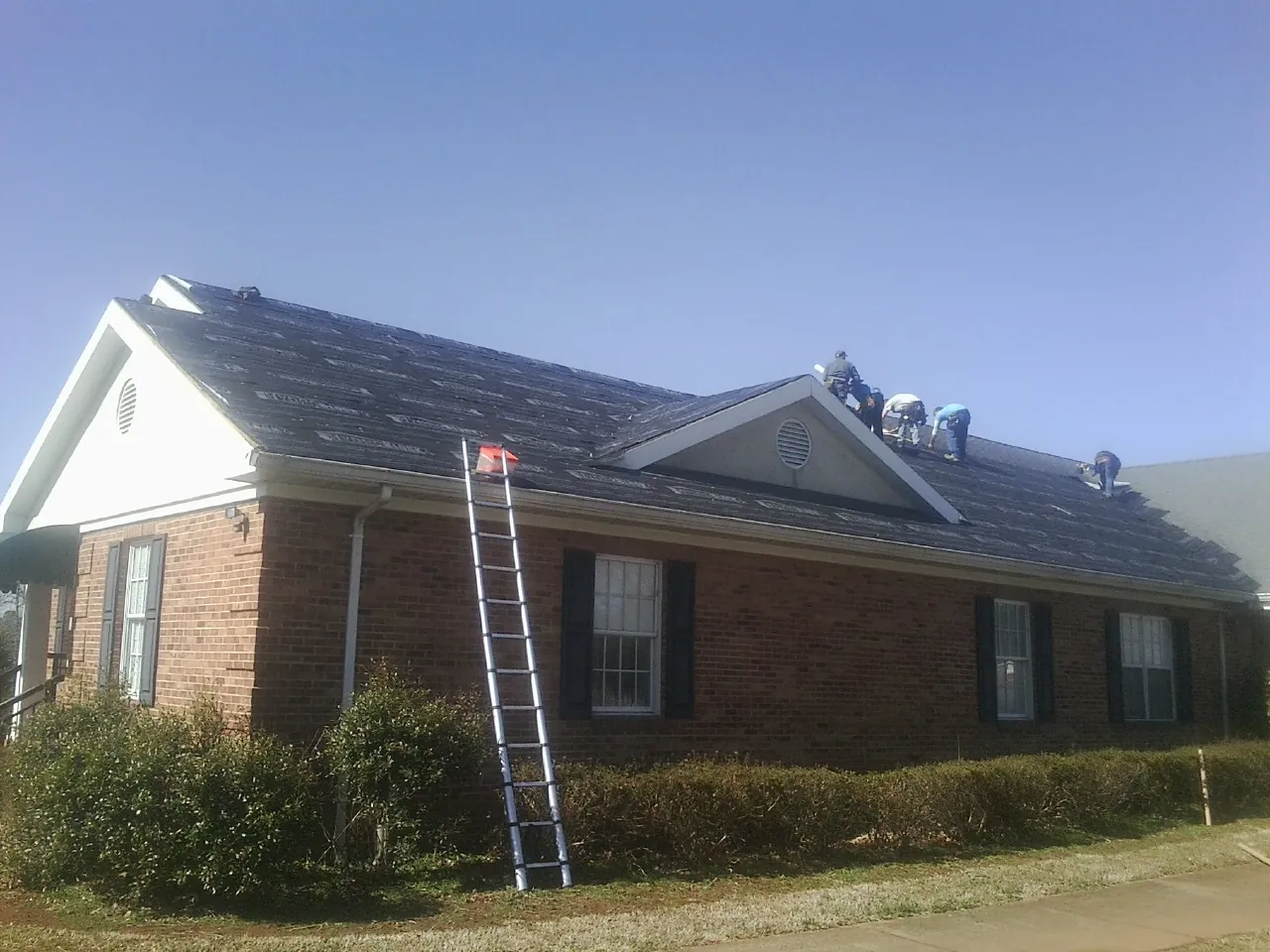 Crew tearing shingles off a brick ranch roof with a ladder set — Metal Roof Repair work by our crew in Saint Johns