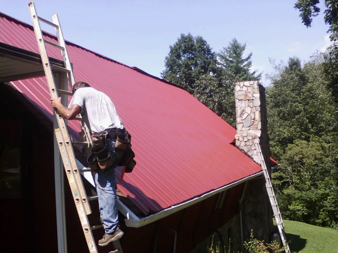 Roofer on a ladder beside a red metal roof and stone chimney — Soffit & Fascia Repair by Roofing Brothers in Saint Johns